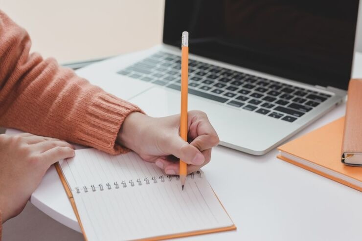 PC sul fondo, primo piano di mano che scrive su quaderno con la matita